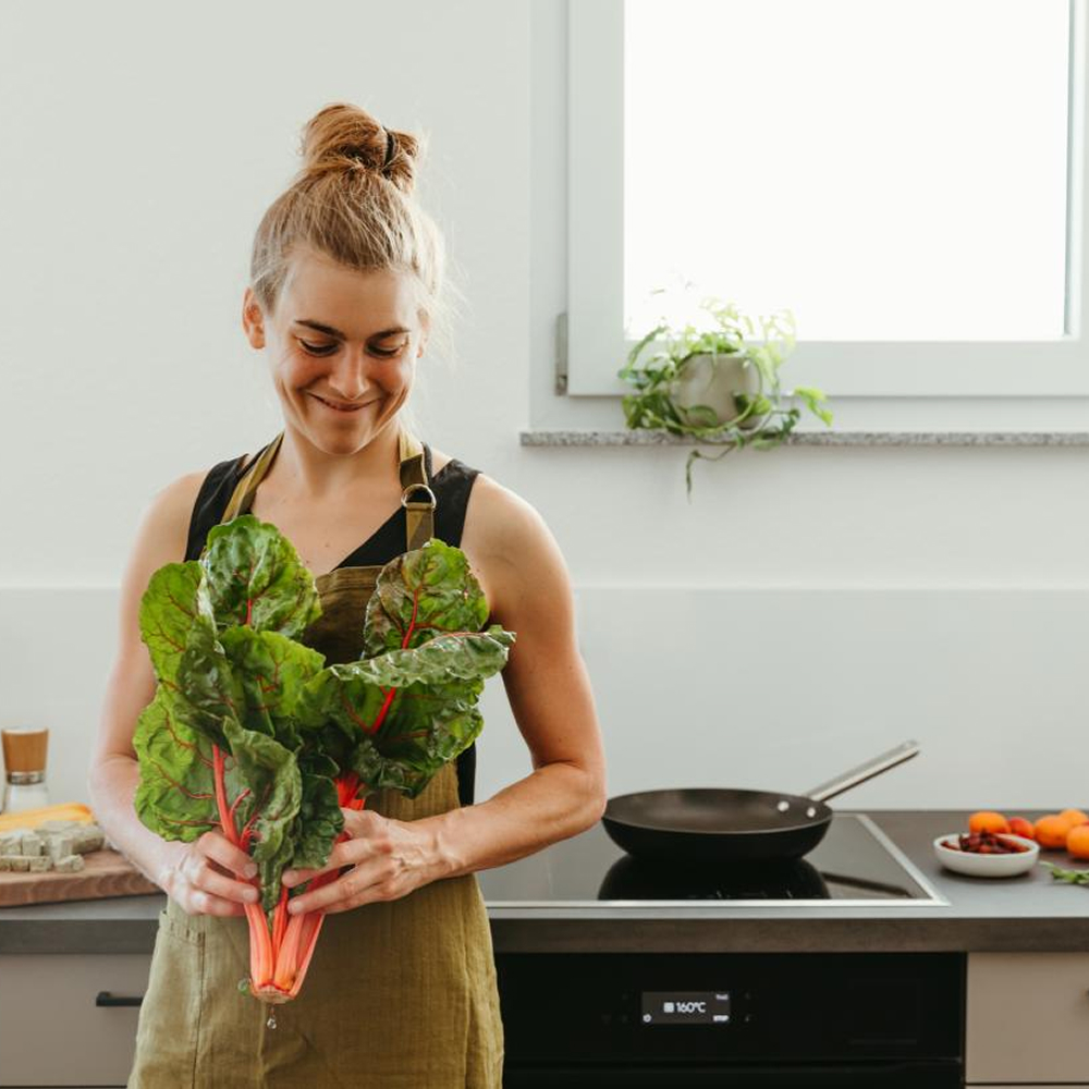 Eine lächelnde Frau mit Dutt und Schürze hält einen großen Bund frischen Mangold mit roten Stielen in den Händen. Sie steht in einer modernen, hellen Küche vor einem Herd mit einer Pfanne.