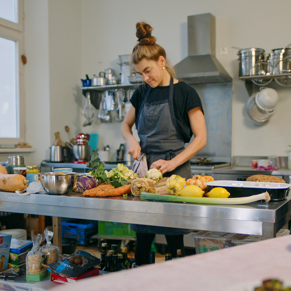 Eine junge Frau mit Dutt und Schürze bereitet in einer hellen Küche frisches Gemüse zu. Sie steht an einem Edelstahltisch voller bunter Zutaten wie Karotten, Blumenkohl und Kürbis und schneidet gerade auf einem Holzbrett.
