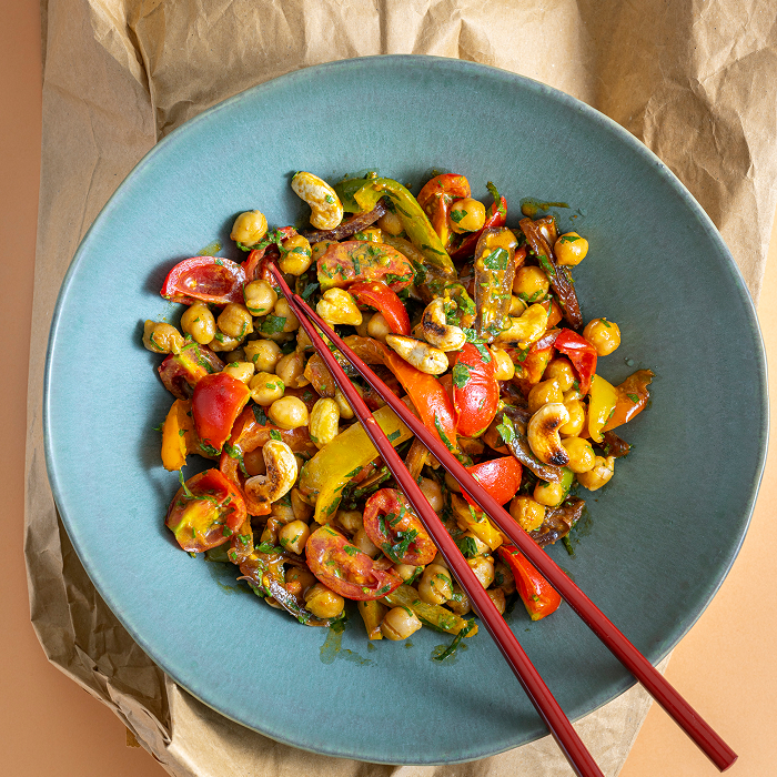 Eine bunte Gemüse-Bowl mit Kichererbsen, halbierten Kirschtomaten, gelben Paprikastreifen und gerösteten Cashewkernen in einer blaugrauen Schüssel. Das Gericht ist mit frischen Kräutern bestreut und zwei rote Essstäbchen liegen quer darüber.