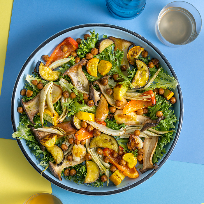 Ein Teller mit einem bunten Salat aus verschiedenen Gemüsesorten, darunter Karotten, Radieschen und grüne Blätter. Der Salat ist mit einer hellen Vinaigrette angemacht und wird von einem Glas Wasser begleitet.