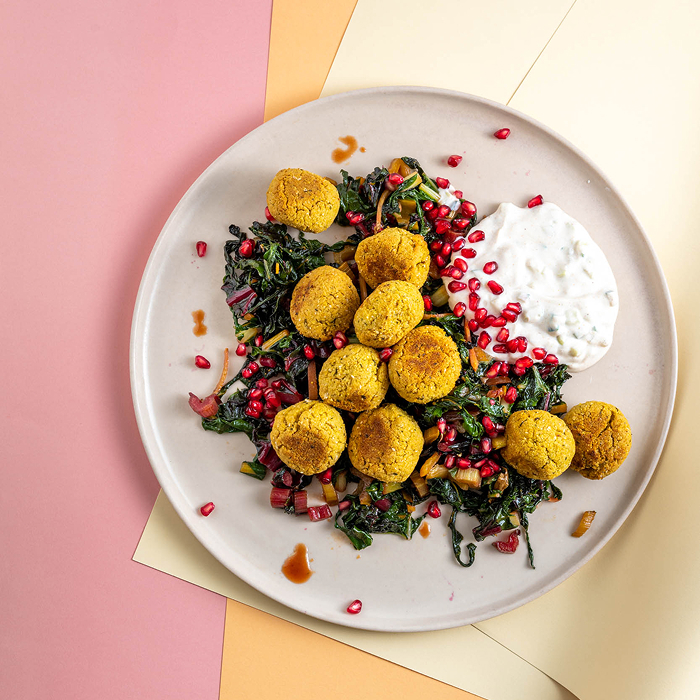 Geriebene gelbe Falafel-Bällchen auf einem Bett aus gedünstetem grünen Blattgemüse und Granatapfelkernen. Serviert auf einem hellen Teller mit einem großen Klecks Kräuterquark, präsentiert vor einem grafischen Hintergrund in Rosa und Pastellgelb.