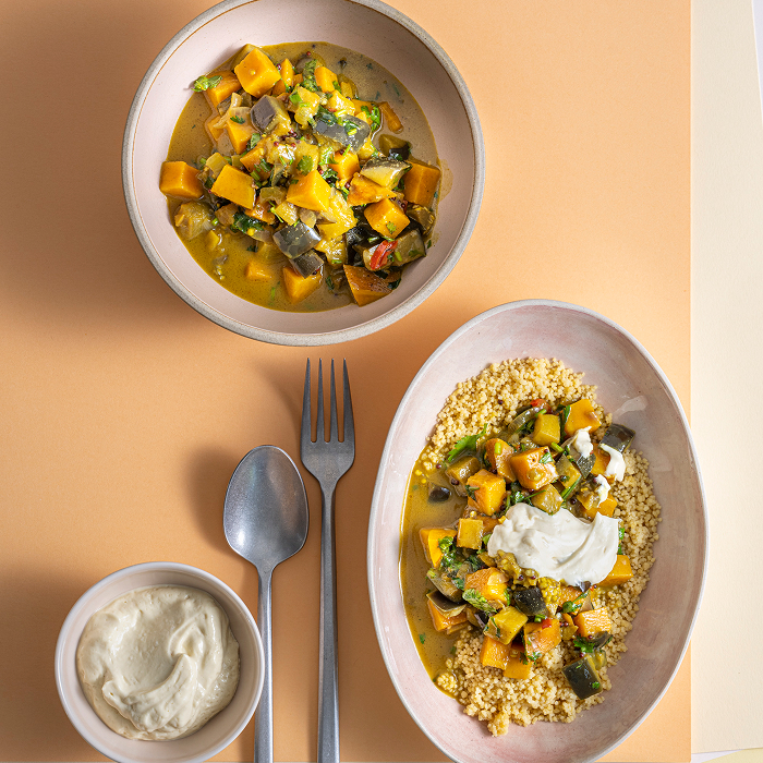 Zwei Keramikschalen mit einem gelben Kürbis-Auberginen-Curry auf pastellfarbenem Hintergrund. Eine Schale enthält nur das Curry, die andere ist zusätzlich mit Couscous und einem Klecks weißer Creme angerichtet. Ein Löffel, eine Gabel und ein Schälchen mit Dip ergänzen das Gedeck.
