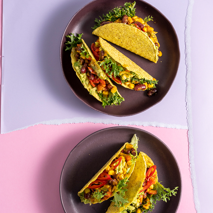 Vier gefüllte Tacos auf zwei dunklen Tellern, angerichtet vor einem zweifarbigen Hintergrund in Rosa und Flieder. Die Tacos sind reichhaltig mit Kidneybohnen, Mais, frischem Salat, Tomaten und Avocado befüllt.