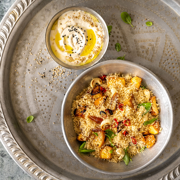 Eine silberne Metallschüssel mit lockerem Couscous, gerösteten Gemüsestücken, Cashewkernen und frischen Basilikumblättern. Daneben steht ein passendes Schälchen mit Hummus, das mit Olivenöl sowie hellem und dunklem Sesam garniert ist, serviert auf einem verzierten Metalltablett.